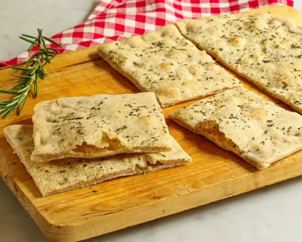 Focaccia senza lievito, croccante e profumata al rosmarino, servita su un tagliere di legno con un tovagliolo a quadri rossi.