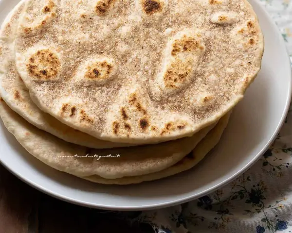 Piatto di piadine senza strutto, dorate e leggermente maculate, impilate su un piatto bianco.