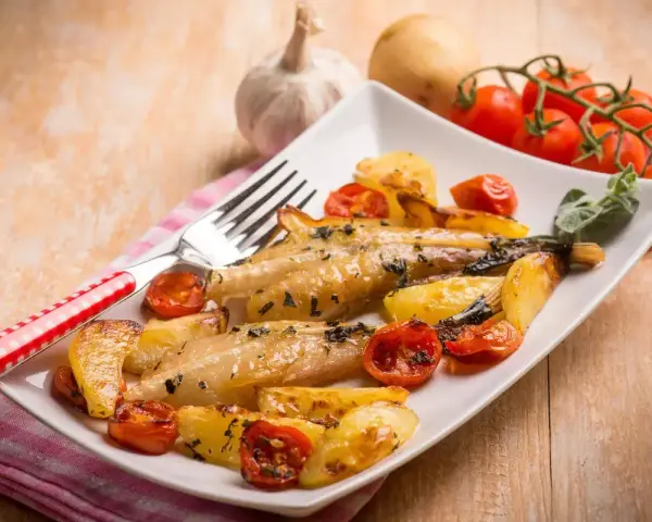 Piatto di coda di rospo al forno con patate arrosto e pomodorini. Un contorno di erbe aromatiche completa il piatto.