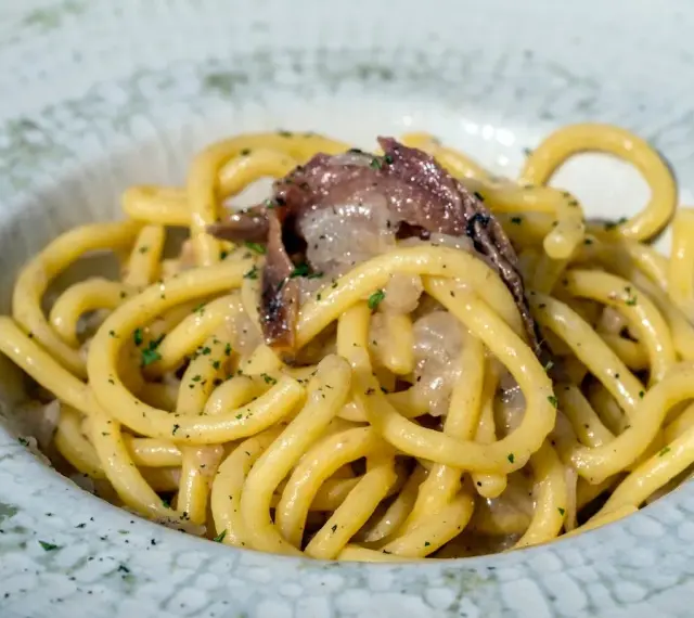 Un piatto di bigoli in salsa, con pasta spessa e condimento a base di acciughe e cipolla, guarnito con prezzemolo tritato.