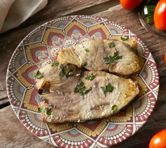Due fette di pesce spada cotto in friggitrice ad aria, guarnite con prezzemolo, su un piatto decorato. Accanto, pomodori, limoni e zucchine.