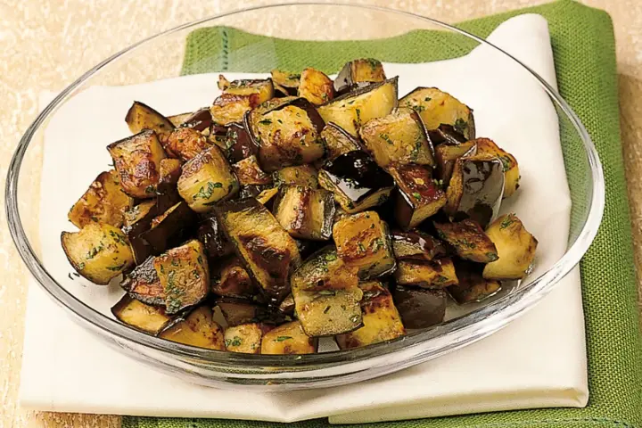 Melanzane a funghetto ricetta della nonna: cubetti dorati e saporiti, conditi con erbe aromatiche, in una ciotola di vetro.