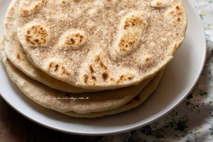 Piatto di piadine senza strutto, dorate e leggermente maculate, impilate su un piatto bianco.