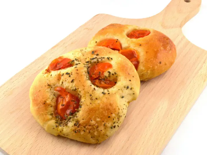 Due focaccine pugliesi dorate, condite con pomodorini e erbe aromatiche, su un tagliere di legno.