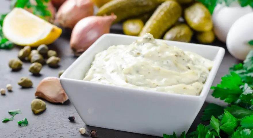Una cremosa salsa remoulade in una ciotola bianca, circondata da ingredienti freschi come limone, capperi, aglio e cetriolini.