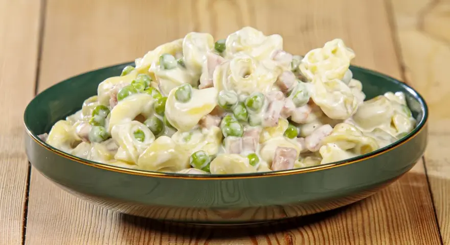 Un piatto invitante di tortellini panna e prosciutto, con piselli verdi, conditi con una cremosa salsa bianca.
