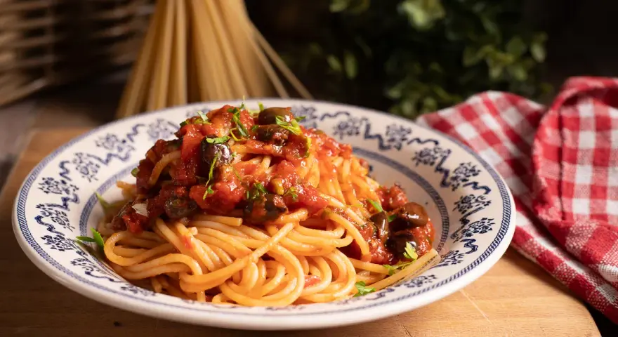 Piatto di spaghetti alla puttanesca, conditi con pomodoro, olive e capperi, guarniti con prezzemolo fresco.