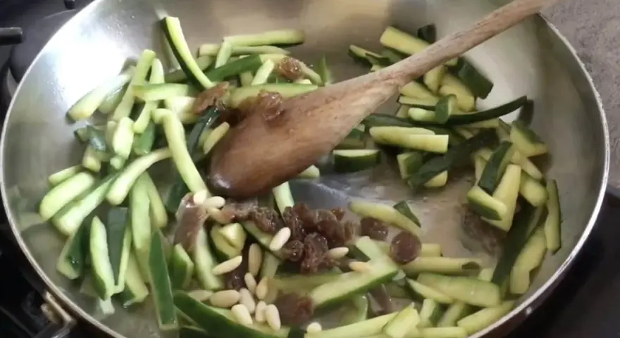 Zucchine in agrodolce veloci in padella con pinoli e uvetta, mescolate con un cucchiaio di legno.