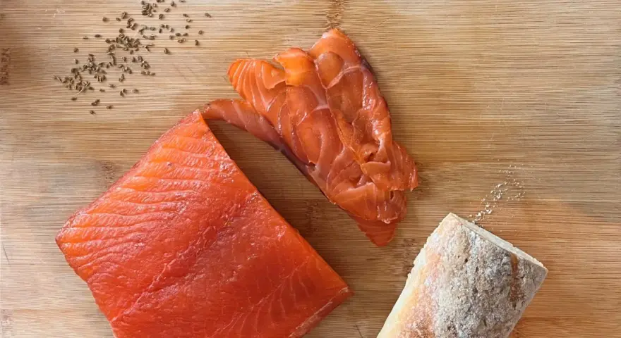 Fetta di salmone marinato, affettato e pane su tagliere di legno. Semi di cumino e sale completano la scena.