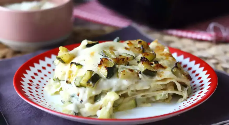 Una fetta di lasagne vegetariane con zucchine e besciamella, dorata al forno, servita su un piatto rosso e bianco.