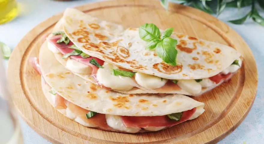 Piadina romagnola ripiena di prosciutto crudo, mozzarella e basilico, servita su un tagliere di legno.