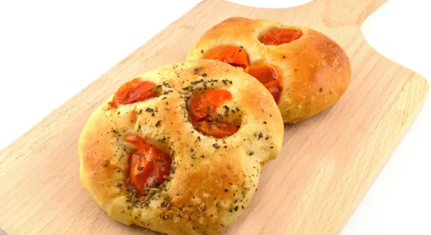Due focaccine pugliesi dorate, condite con pomodorini e erbe aromatiche, su un tagliere di legno.