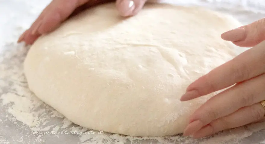 Mani che lavorano un soffice impasto pizza su un piano infarinato, pronte per essere stese.