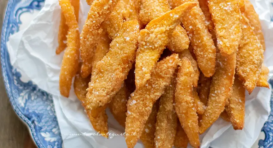 Un mucchio di bastoncini dorati di zucca fritta impanata, croccanti e salati, serviti su carta bianca in un piatto blu e bianco.