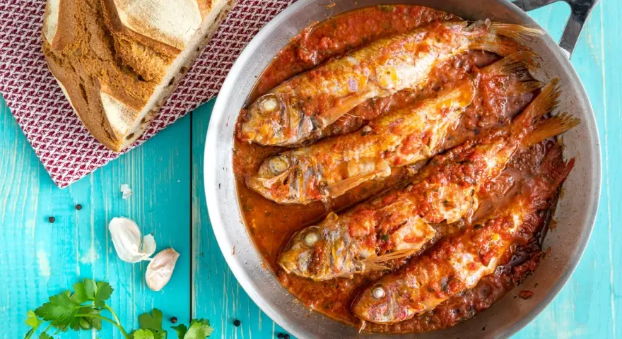 Triglie alla livornese in padella, con pane rustico, aglio e prezzemolo su tavolo blu.