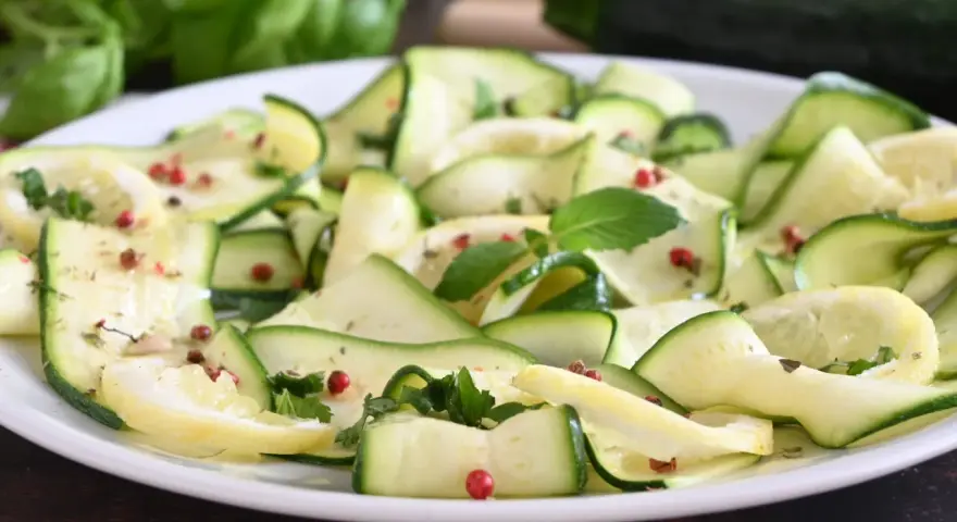 Zucchine marinate a fettine sottili, condite con erbe aromatiche e pepe rosa, su un piatto bianco.