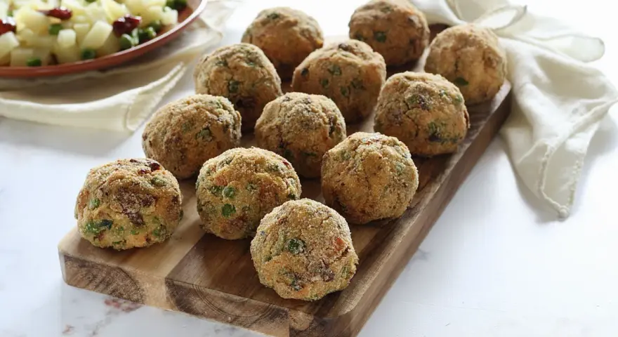Deliziose polpette di verdure dorate e croccanti, disposte su un tagliere di legno. Un contorno di patate e piselli sullo sfondo.