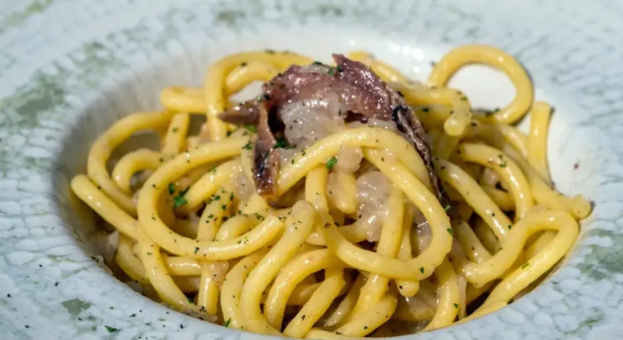Un piatto di bigoli in salsa, con pasta spessa e condimento a base di acciughe e cipolla, guarnito con prezzemolo tritato.