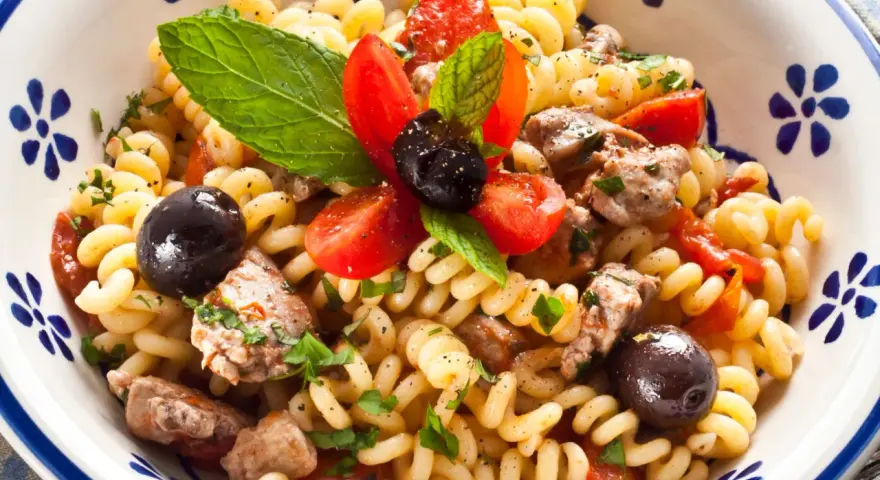 Fusilli con pesce spada, pomodorini, olive nere e menta fresca in una ciotola decorata.