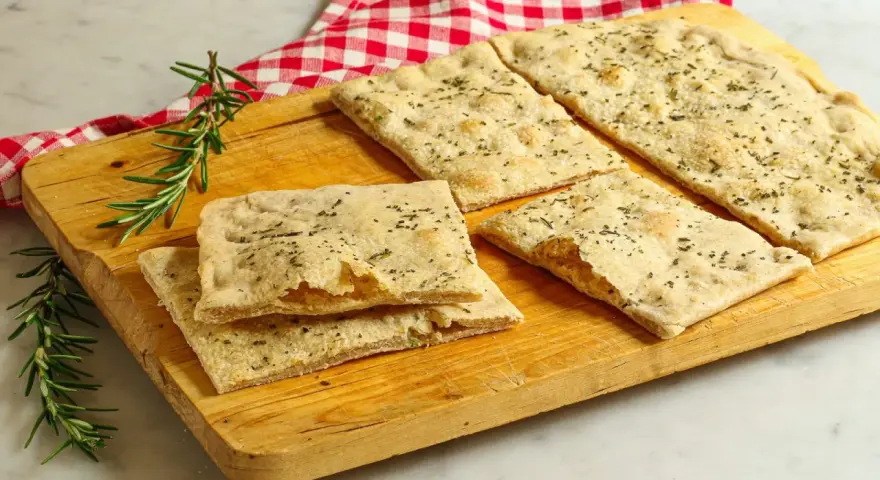 Focaccia senza lievito, croccante e profumata al rosmarino, servita su un tagliere di legno con un tovagliolo a quadri rossi.