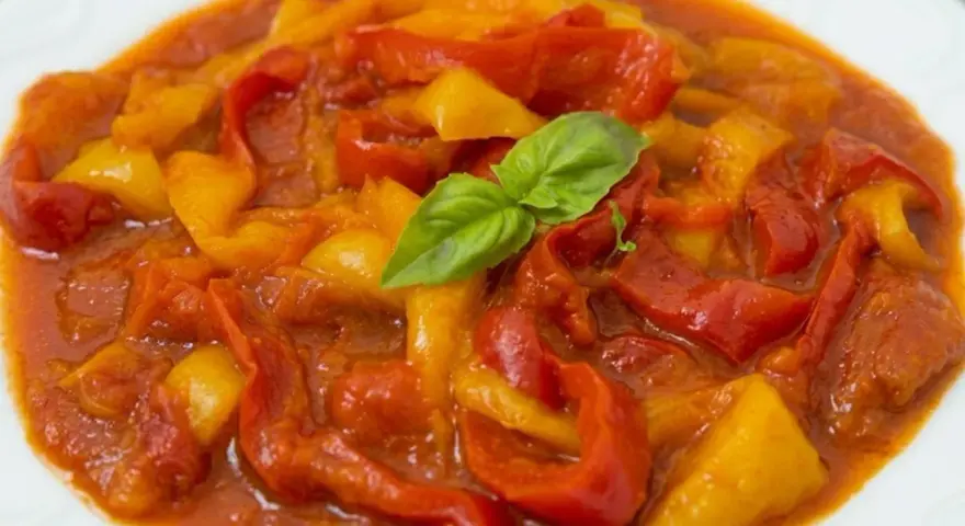 Un piatto di peperonata ricetta, con peperoni rossi e gialli cotti in salsa, guarnito con basilico fresco.
