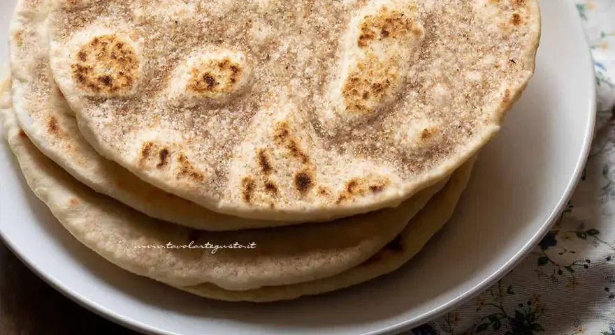 Piatto di piadine senza strutto, dorate e leggermente maculate, impilate su un piatto bianco.