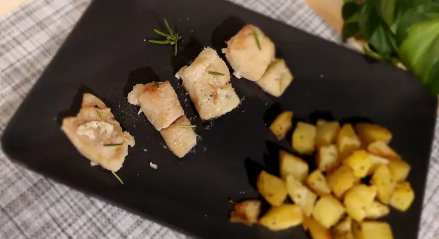 Pezzi di baccalà al forno con rosmarino e patate arrosto su piatto nero.
