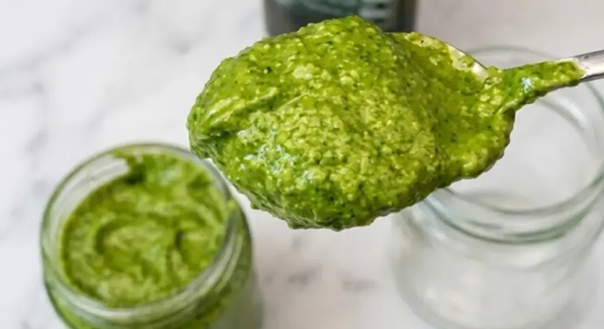 Cucchiaio pieno di pesto senza pinoli, cremoso e verde brillante, accanto a un barattolo.