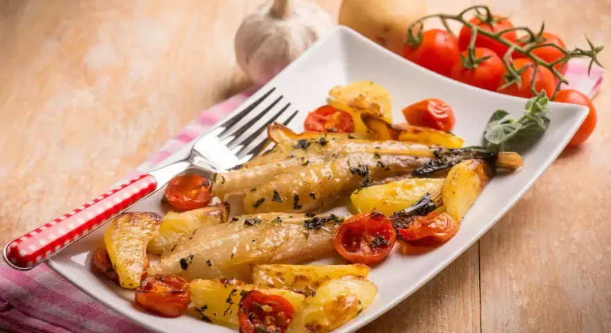 Piatto di coda di rospo al forno con patate arrosto e pomodorini. Un contorno di erbe aromatiche completa il piatto.