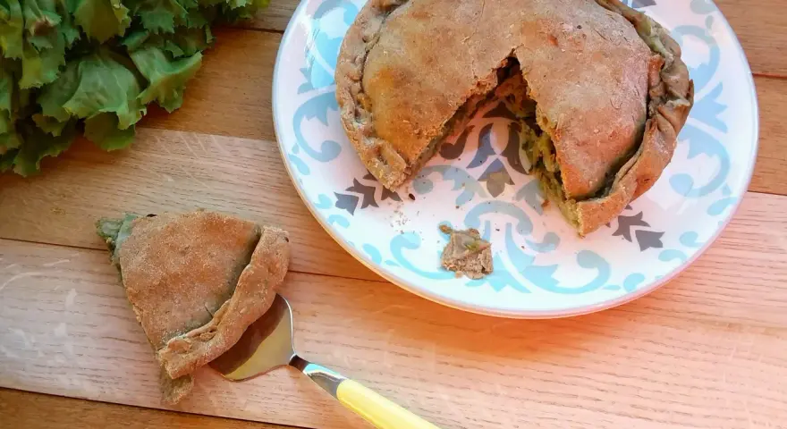 Torta salata con scarola, acciughe e mozzarella, appena sfornata, con una fetta servita su un piatto decorato.