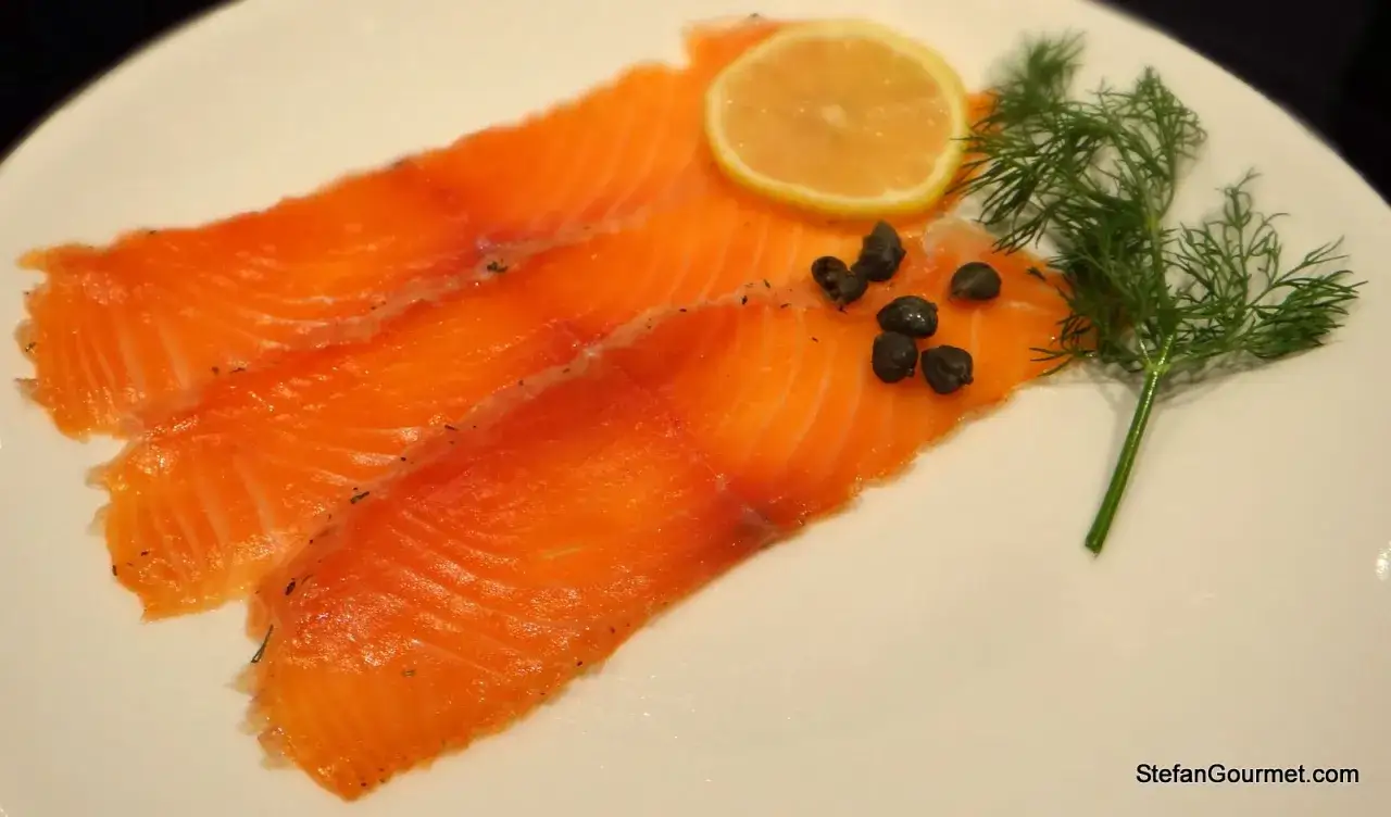 Fette di salmone marinato su piatto bianco, guarnite con capperi, una fetta di limone e un rametto di aneto.