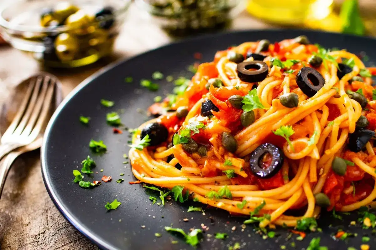 Un piatto di spaghetti alla puttanesca, con pomodoro, olive nere, capperi e prezzemolo fresco, pronto per essere gustato.