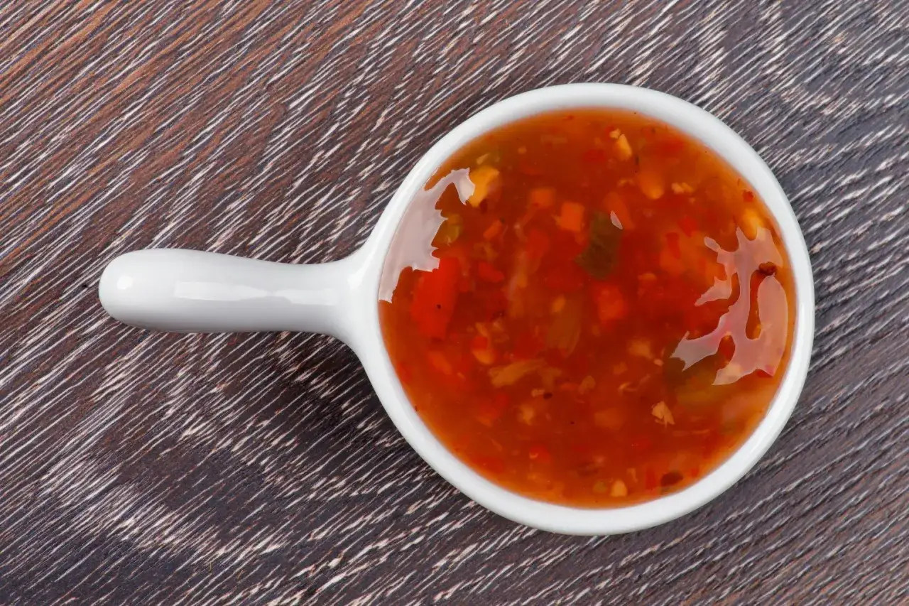 Una ciotolina bianca contiene una vibrante salsa agrodolce, con pezzetti di peperone e spezie.