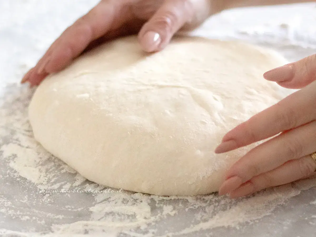 Mani che lavorano un soffice impasto pizza su un piano infarinato, pronte per essere stese e condite.