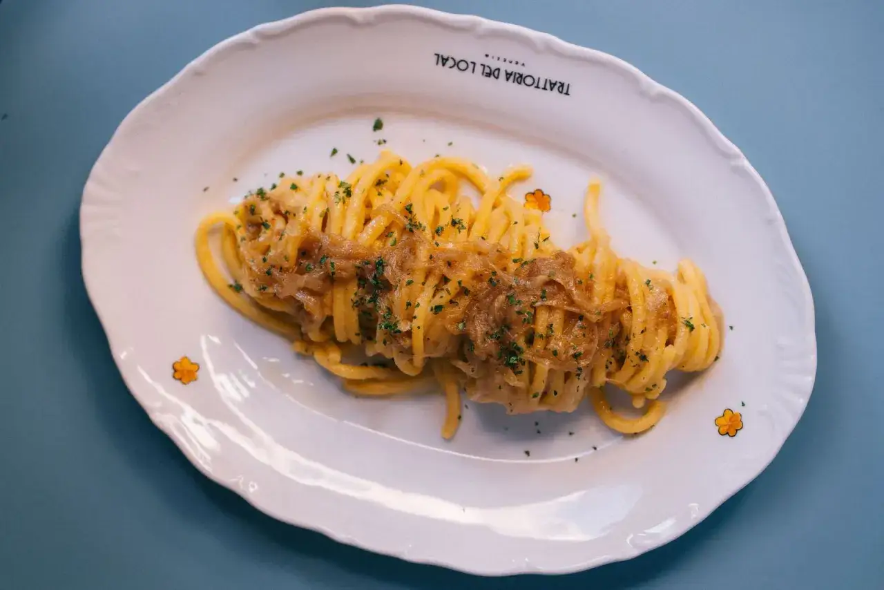 Un piatto di bigoli in salsa, pasta spessa condita con cipolle caramellate e prezzemolo tritato, servito su un piatto bianco con decorazioni floreali.