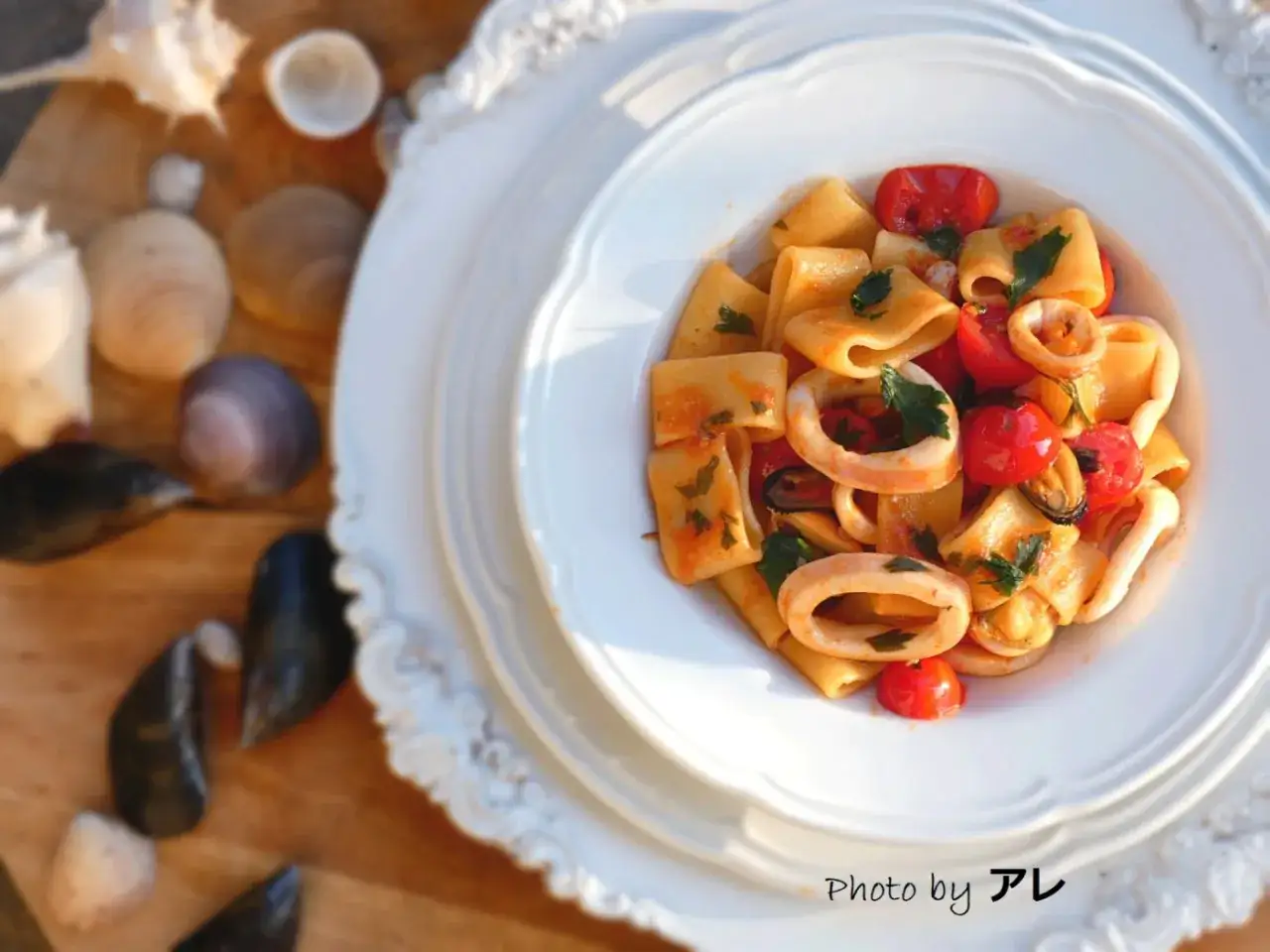 Piatto di calamarata allo scoglio con calamari, cozze, pomodorini e prezzemolo, su un tavolo con conchiglie e cozze.