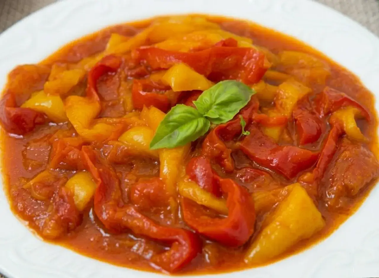 Un piatto invitante di peperonata ricetta, con peperoni rossi e gialli cotti lentamente in un sugo saporito, guarnito con una foglia di basilico fresco.
