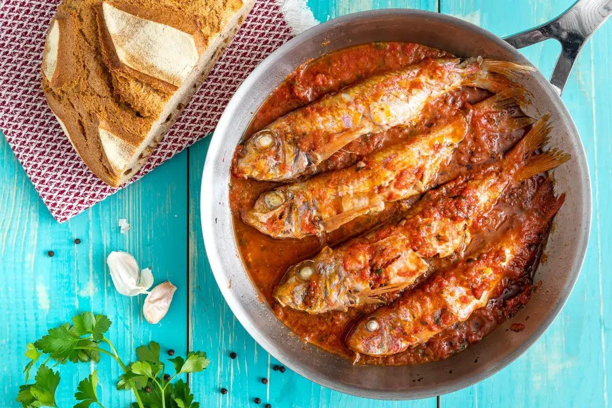 Triglie alla livornese in padella, con pane rustico, aglio e prezzemolo su tavolo azzurro.