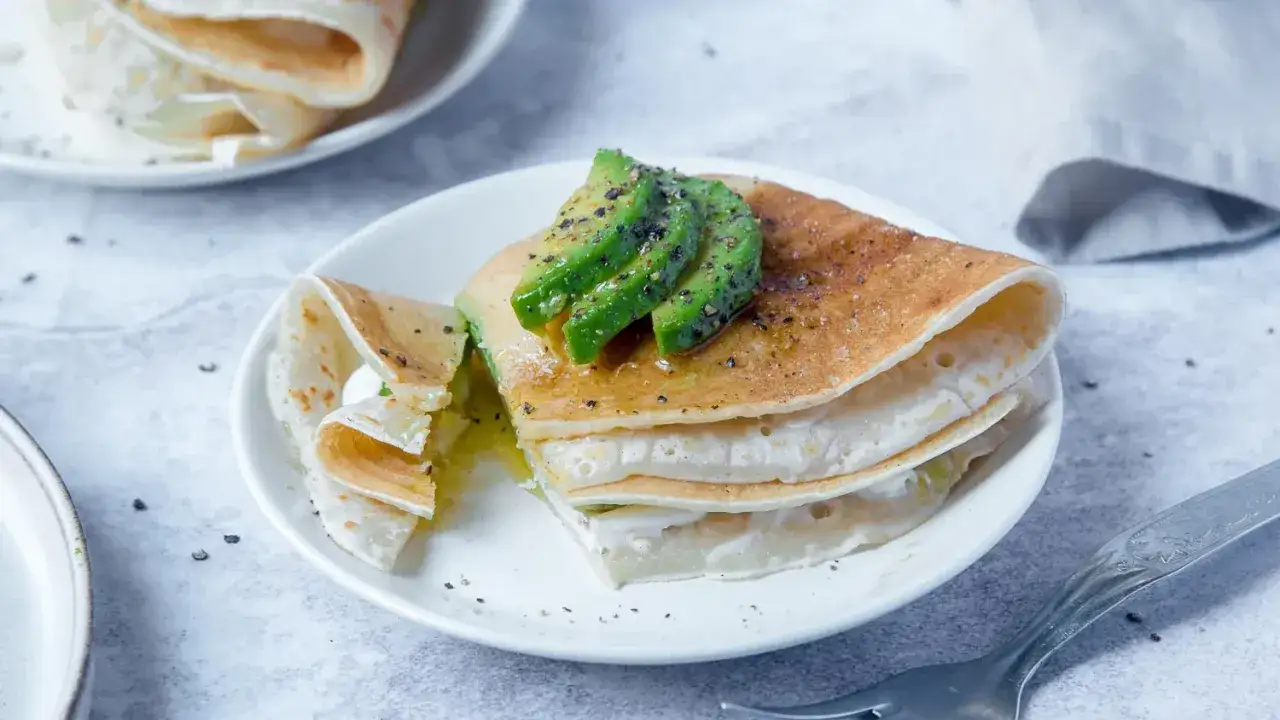 Deliziose crepes senza uova, impilate e guarnite con fette di avocado fresco e un filo d'olio.