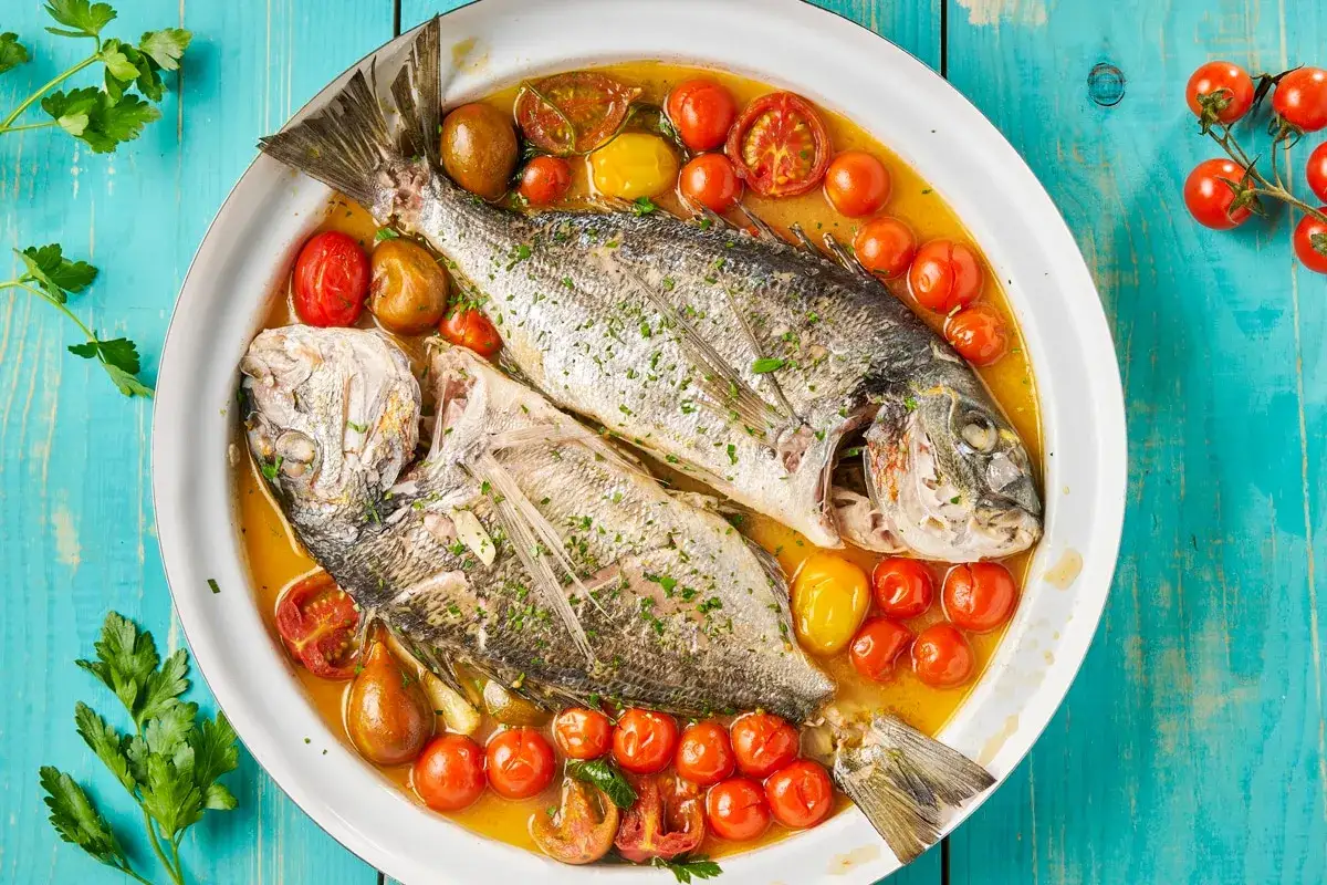 Orata all'acqua pazza, due pesci interi cotti in un brodo saporito con pomodorini e erbe aromatiche, serviti in un piatto bianco su un tavolo di legno blu.
