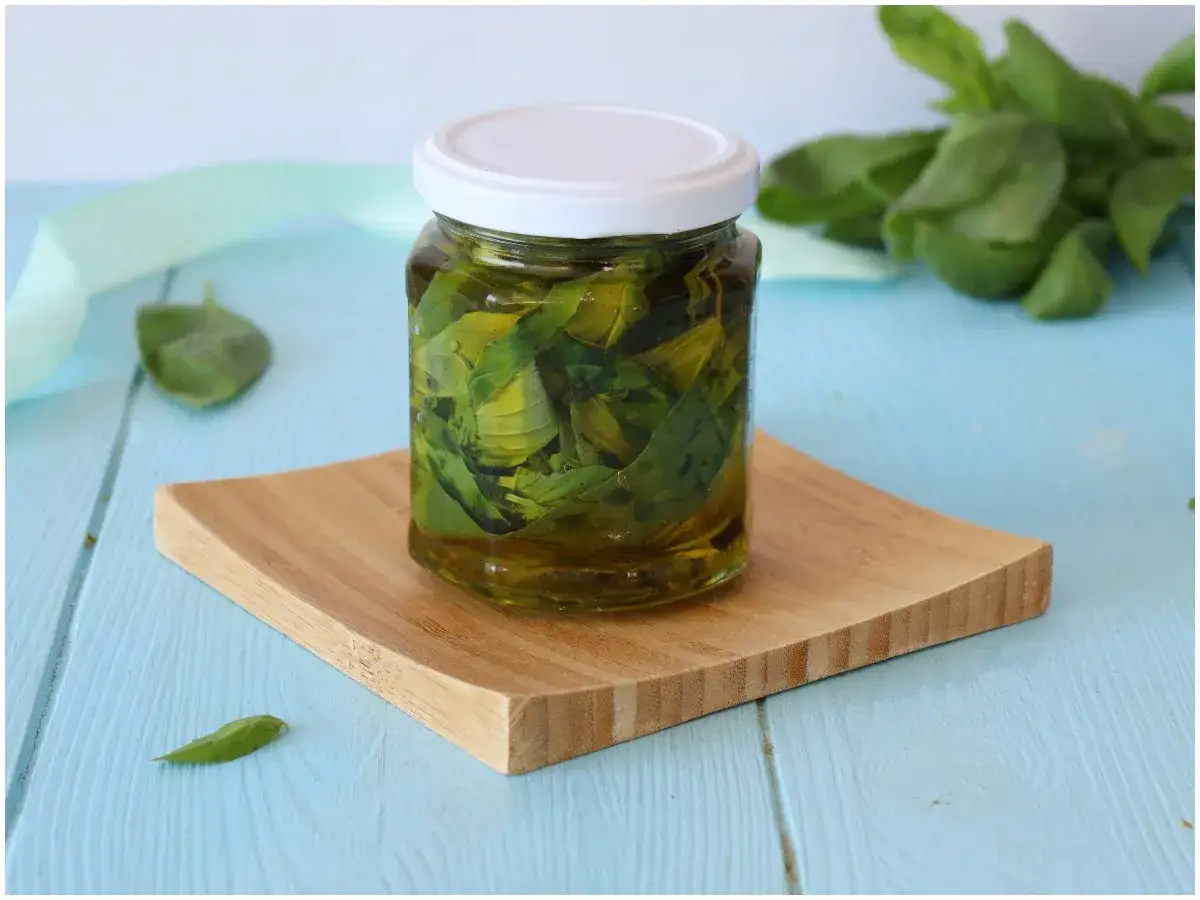 Un vasetto di basilico sott'olio, con foglie fresche sullo sfondo, su un tagliere di legno.