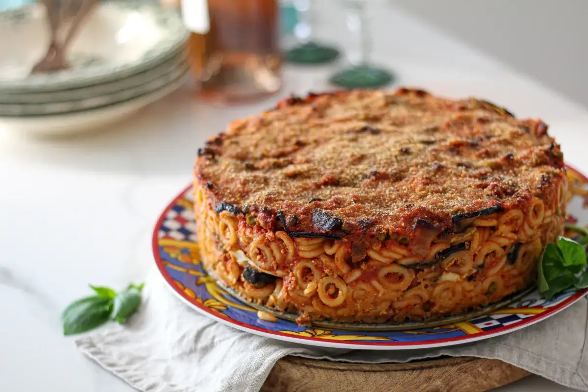 Un tortino rotondo di anelletti alla siciliana, con melanzane e sugo, cotto al forno e servito su un piatto decorato.