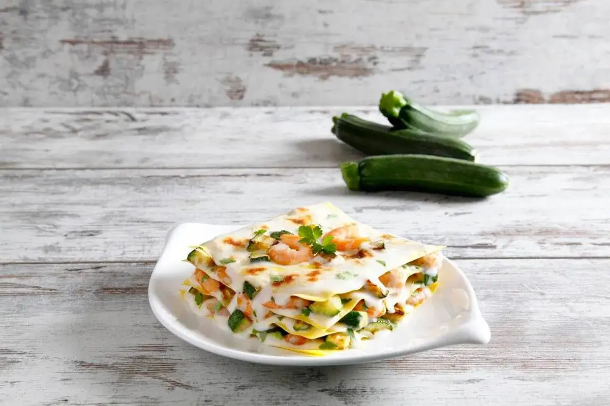 Deliziosa lasagna estiva con zucchine e gamberetti, guarnita con prezzemolo fresco. Sullo sfondo, zucchine fresche su un tavolo di legno.