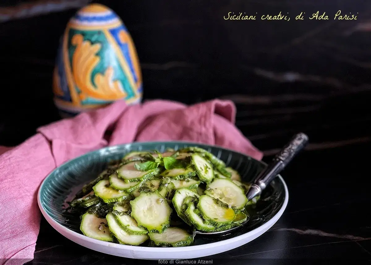 Zucchine marinate fresche e profumate, condite con erbe aromatiche e olio, servite in un piatto verde su un tovagliolo rosa.