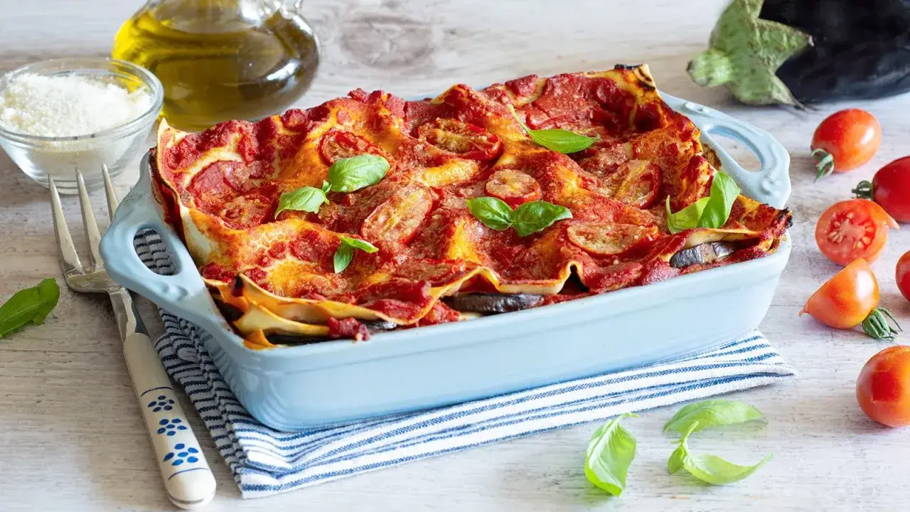 Lasagna di melanzane appena sfornata, dorata e fumante, guarnita con basilico fresco. Accanto, pomodorini, olio d'oliva e parmigiano.