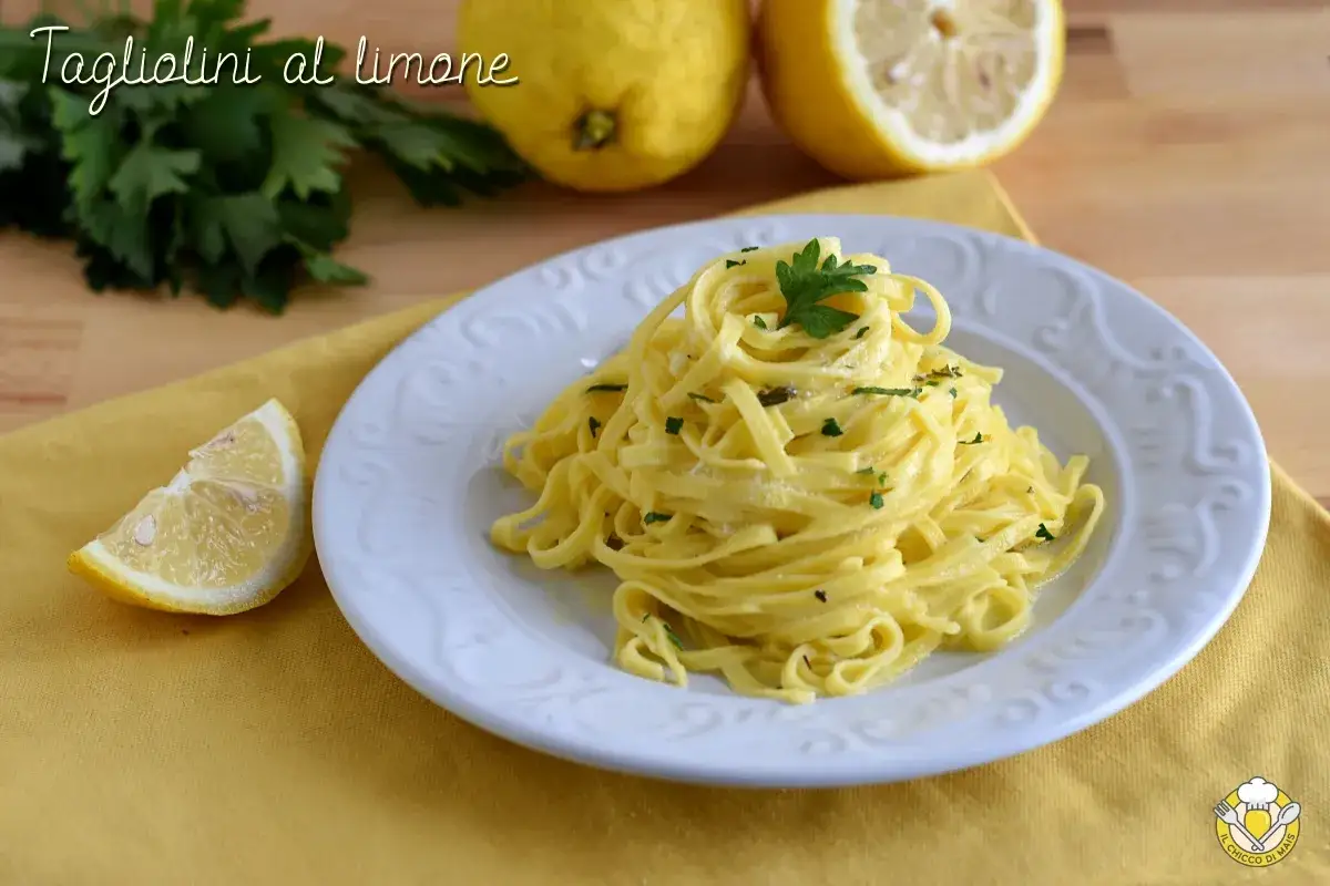 Un piatto di tagliolini al limone, guarnito con prezzemolo fresco, accompagnato da spicchi di limone e limoni interi sullo sfondo.