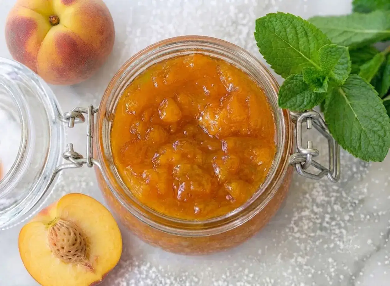 Vasetto di marmellata di pesche fatto in casa, con pesche fresche e foglie di menta.