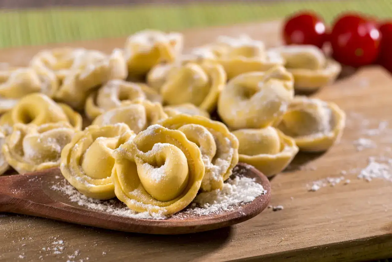 Tortellini fatti in casa, pronti per essere cucinati, spolverati di farina su un tagliere di legno, con pomodorini sullo sfondo.