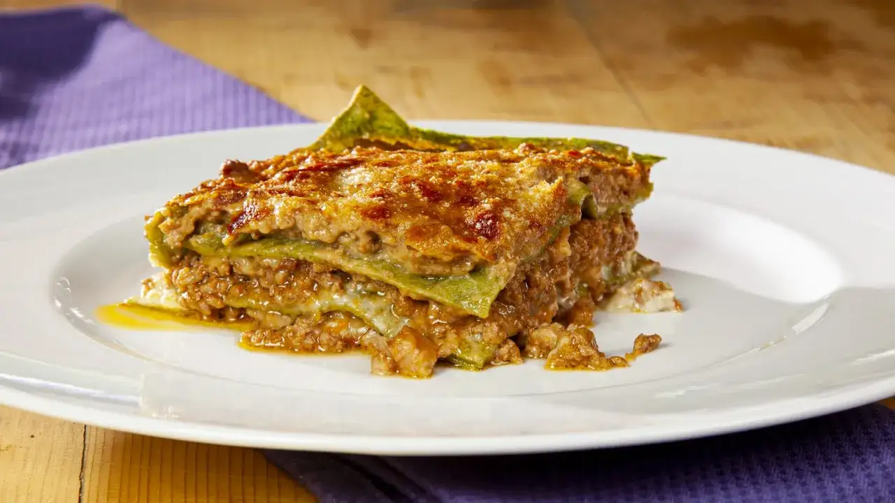 Una fetta di lasagna alla bolognese, con strati di pasta verde, ragù e formaggio fuso, servita su un piatto bianco.