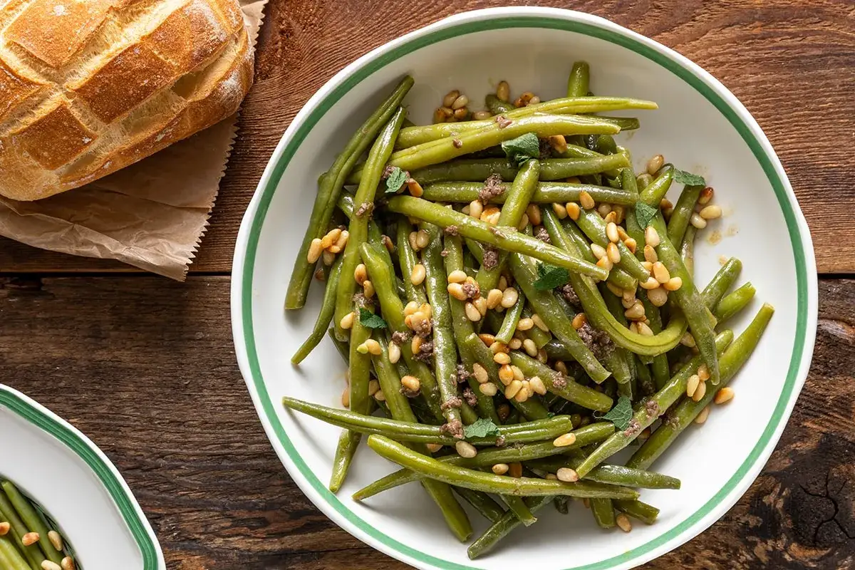 Fagiolini lessi conditi con pinoli tostati e un tocco di erbe aromatiche, serviti in una ciotola bianca con bordo verde. Accanto, una pagnotta rustica.